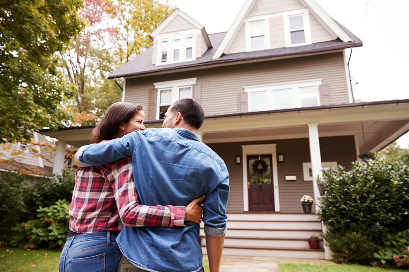 Low Down Payment Purchase Options 1 A couple stands arm-in-arm, looking at a two-story house with a porch and steps, surrounded by greenery and trees, on a sunny day.