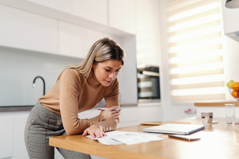 A woman stands in a modern kitchen, leaning over a wooden counter while reviewing documents. A tablet, glass of water, and bowl of fruit are also on the counter beside her.