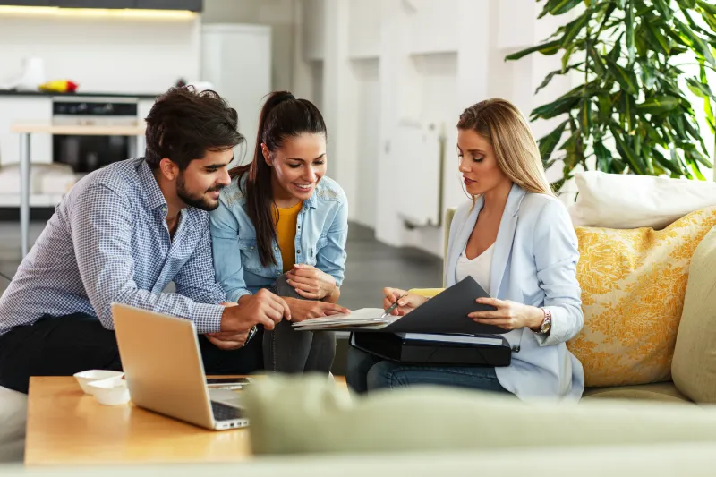 HELOC Home Loan 1 A professional woman shows documents to a smiling couple seated on a sofa in a modern, bright living room. A laptop and coffee cup are on the table, and a large plant is in the background.
